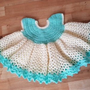 K&ouml;nnte beinhalten: Ein geh&auml;keltes Babykleid in Wei&szlig; und T&uuml;rkis. Das Kleid hat einen gewellten Saum und einen runden Ausschnitt.