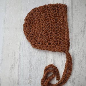 May include: A brown crocheted baby bonnet with a long tie. The bonnet is made with a simple stitch pattern and has a rounded shape.