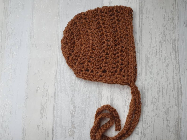 May include: A brown crocheted baby bonnet with a long tie. The bonnet is made with a simple stitch pattern and has a rounded shape.