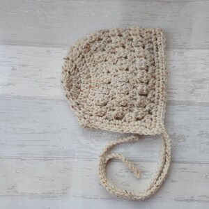 K&ouml;nnte beinhalten: Eine geh&auml;kelte Babyhaube in Cremefarbe mit einer Schleife. Die Haube ist mit einem strukturierten Stich gearbeitet und hat ein schlichtes Design.