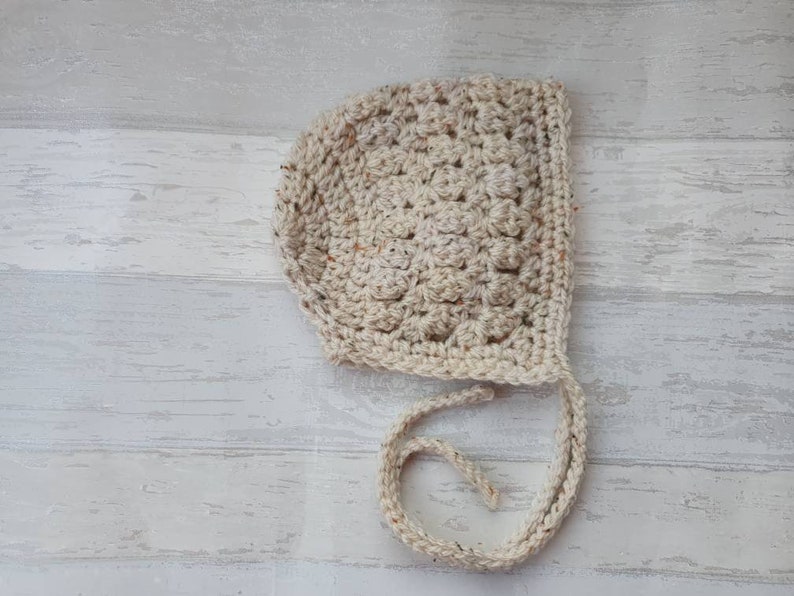 K&ouml;nnte beinhalten: Eine geh&auml;kelte Babyhaube in Cremefarbe mit einer Schleife. Die Haube ist mit einem strukturierten Stich gearbeitet und hat ein schlichtes Design.