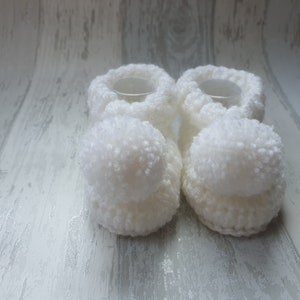 May include: A pair of white crocheted baby booties with fluffy pom-poms on the toes.