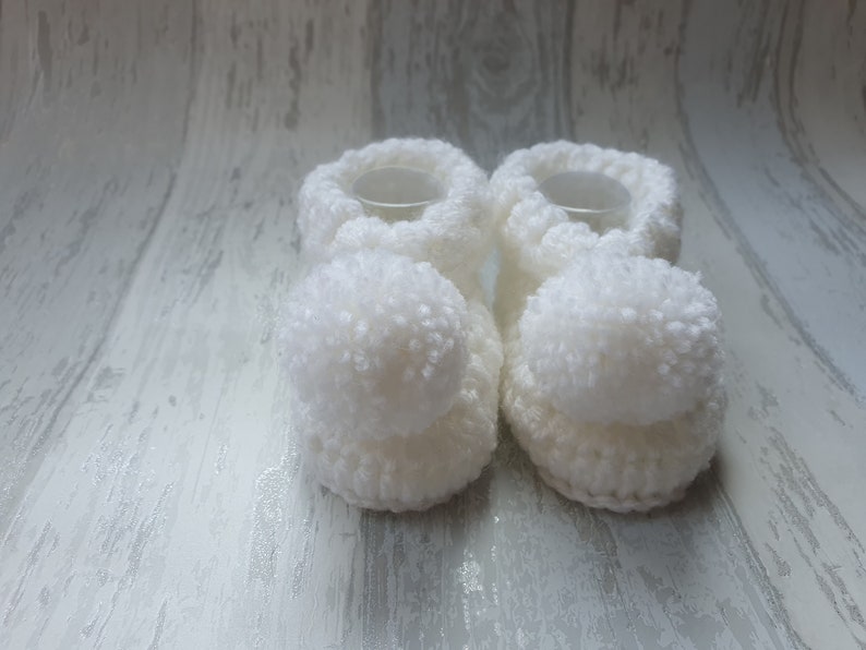 May include: A pair of white crocheted baby booties with fluffy pom-poms on the toes.