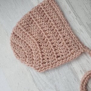 May include: A crocheted baby bonnet in a pale pink color with a long tie. The bonnet is made with a simple stitch pattern and has a soft, delicate look.