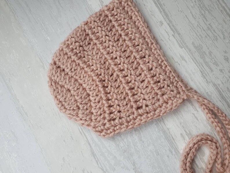 May include: A crocheted baby bonnet in a pale pink color with a long tie. The bonnet is made with a simple stitch pattern and has a soft, delicate look.