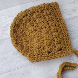 K&ouml;nnte beinhalten: Eine gelbe, geh&auml;kelte Babyhaube mit langem Band. Die Haube ist mit einem strukturierten Stichmuster gearbeitet.