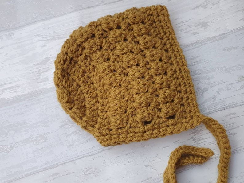 K&ouml;nnte beinhalten: Eine gelbe, geh&auml;kelte Babyhaube mit langem Band. Die Haube ist mit einem strukturierten Stichmuster gearbeitet.