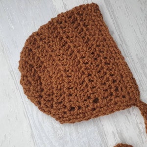 May include: A brown crocheted baby bonnet with a long tie. The bonnet is made with a simple stitch pattern and has a soft, cozy feel.