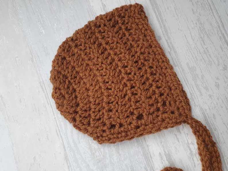 May include: A brown crocheted baby bonnet with a long tie. The bonnet is made with a simple stitch pattern and has a soft, cozy feel.
