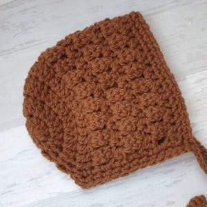 K&ouml;nnte beinhalten: Eine braune geh&auml;kelte Babyhaube mit einer gebundenen Schnur. Die Haube ist mit einem strukturierten Stichmuster gearbeitet.