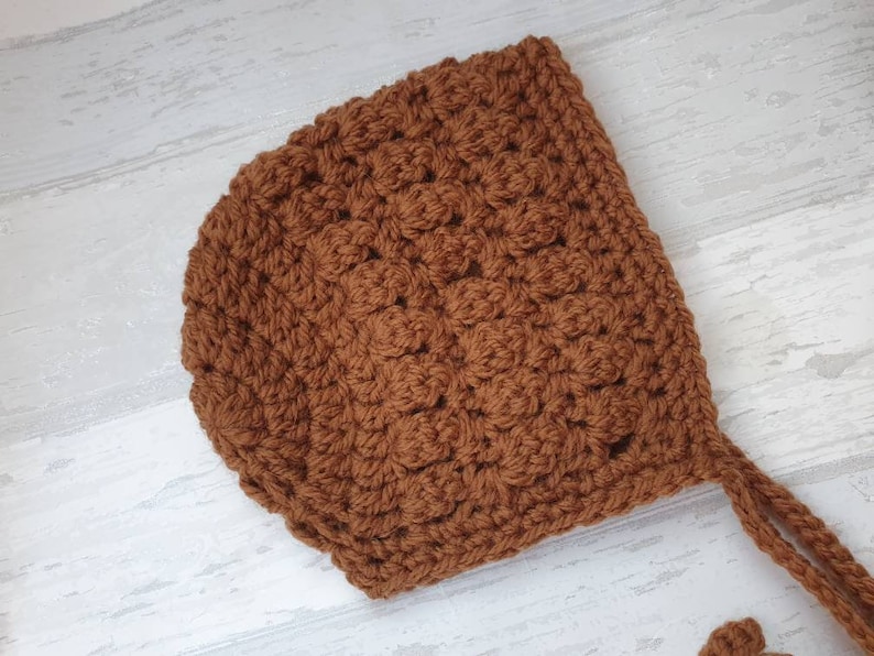 K&ouml;nnte beinhalten: Eine braune geh&auml;kelte Babyhaube mit einer gebundenen Schnur. Die Haube ist mit einem strukturierten Stichmuster gearbeitet.