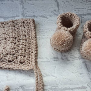 May include: A set of crocheted baby booties and a bonnet in a light brown color. The booties have pom-poms on the toes.