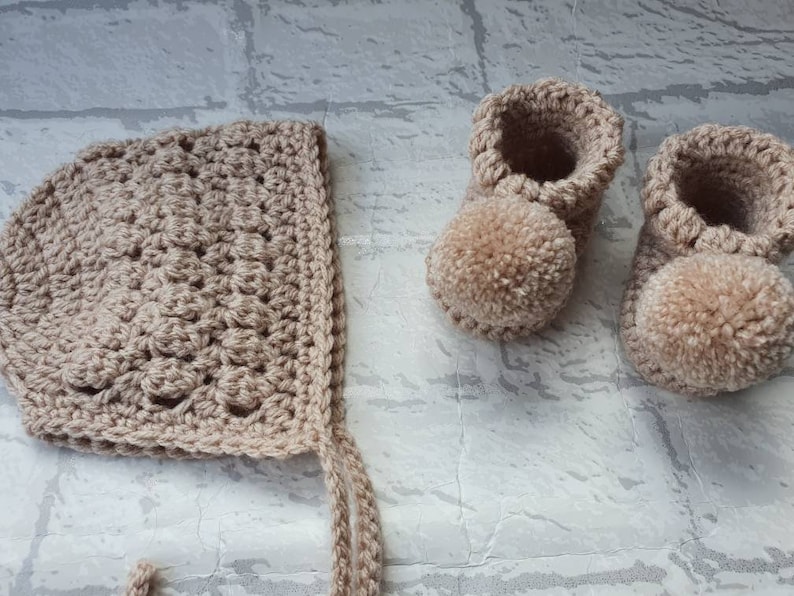May include: A set of crocheted baby booties and a bonnet in a light brown color. The booties have pom-poms on the toes.