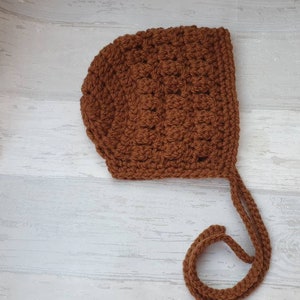 K&ouml;nnte beinhalten: Eine braune geh&auml;kelte Babyhaube mit langem Band. Die Haube ist mit einem strukturierten Stich gearbeitet und hat ein schlichtes Design.