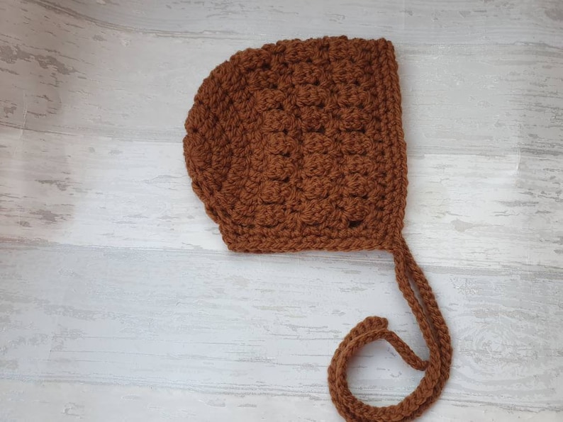 K&ouml;nnte beinhalten: Eine braune geh&auml;kelte Babyhaube mit langem Band. Die Haube ist mit einem strukturierten Stich gearbeitet und hat ein schlichtes Design.