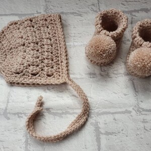 May include: A set of crocheted baby booties and a bonnet in a light beige color. The booties have pom-poms on the toes.