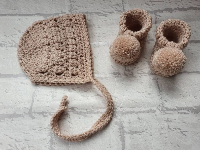 May include: A set of crocheted baby booties and a bonnet in a light beige color. The booties have pom-poms on the toes.