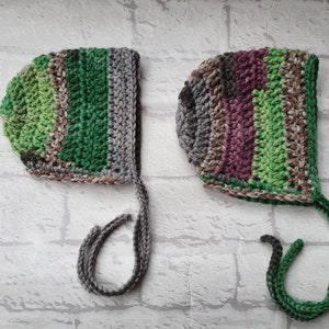 May include: Two crocheted baby bonnets with ties. One bonnet is green, grey, and brown. The other bonnet is green, brown, and purple.