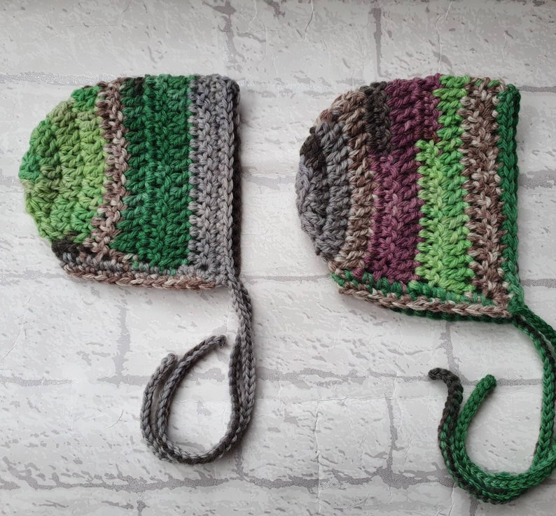 May include: Two crocheted baby bonnets with ties. One bonnet is green, grey, and brown. The other bonnet is green, brown, and purple.