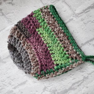 May include: A crocheted baby bonnet with a striped pattern in shades of green, brown, and purple. The bonnet has a tie at the back.