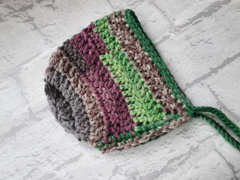 May include: A crocheted baby bonnet with a striped pattern in shades of green, brown, and purple. The bonnet has a tie at the back.