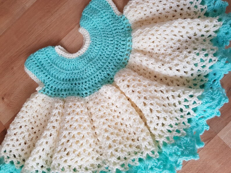 K&ouml;nnte beinhalten: Ein geh&auml;keltes Babykleid in Wei&szlig; und T&uuml;rkis. Das Kleid hat einen R&uuml;schenrock und einen runden Ausschnitt.