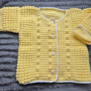 May include: A yellow crocheted cardigan with white trim and buttons. The cardigan has a textured stitch pattern.