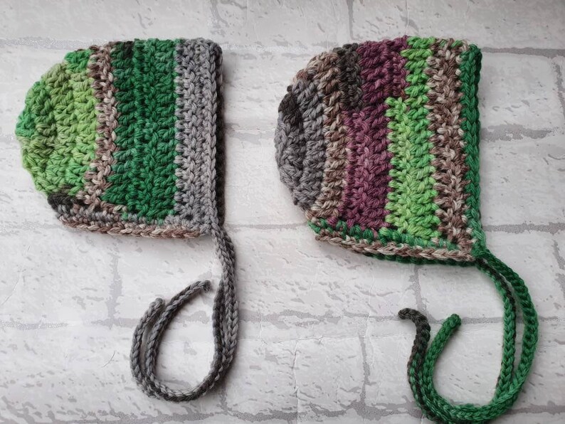 May include: Two crocheted baby bonnets with ties. One is green, brown, and grey. The other is purple, brown, and green.