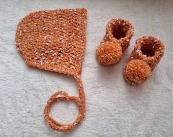 May include: A set of crocheted baby booties and a matching bonnet in a light orange and white color. The booties have pom-poms on the toes.