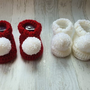 May include: Two pairs of crocheted baby booties with pom-poms. One pair is red and the other is white.