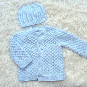 May include: A light blue knitted baby cardigan with a matching hat. The cardigan has a button closure and a textured stitch pattern.