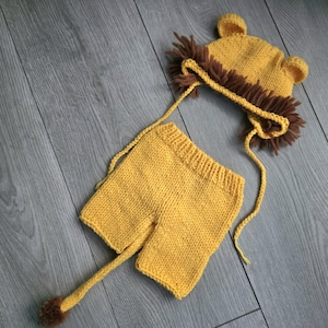May include: A knitted baby outfit designed as a lion. The set features a yellow hat with brown ears and a mane, and matching yellow shorts with a brown tail. The outfit is made of soft yarn.