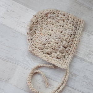 K&ouml;nnte beinhalten: Eine geh&auml;kelte Babyhaube in Cremefarbe mit einem strukturierten Stichmuster. Die Haube hat einen Bindegurt an der R&uuml;ckseite.
