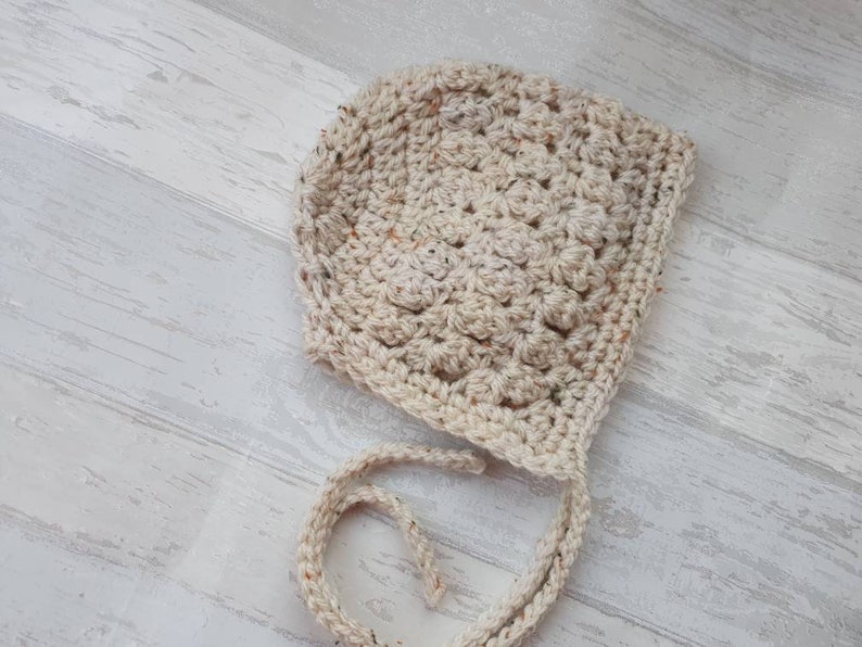 K&ouml;nnte beinhalten: Eine geh&auml;kelte Babyhaube in Cremefarbe mit einem strukturierten Stichmuster. Die Haube hat einen Bindegurt an der R&uuml;ckseite.