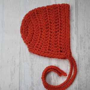 May include: A red crocheted baby bonnet with a long tie. The bonnet is made with a simple stitch pattern and has a rounded shape.