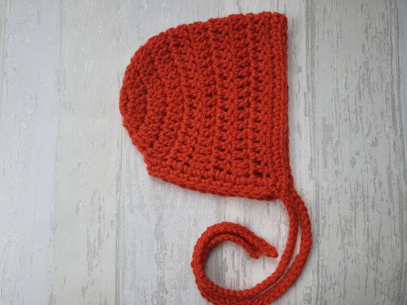 May include: A red crocheted baby bonnet with a long tie. The bonnet is made with a simple stitch pattern and has a rounded shape.