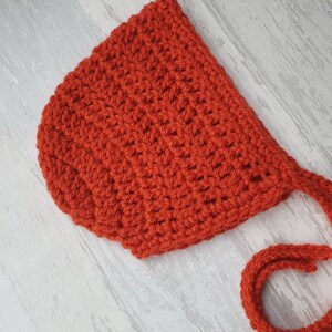 May include: A red crocheted baby bonnet with a long tie. The bonnet is made with a simple stitch pattern and has a soft, cozy feel.