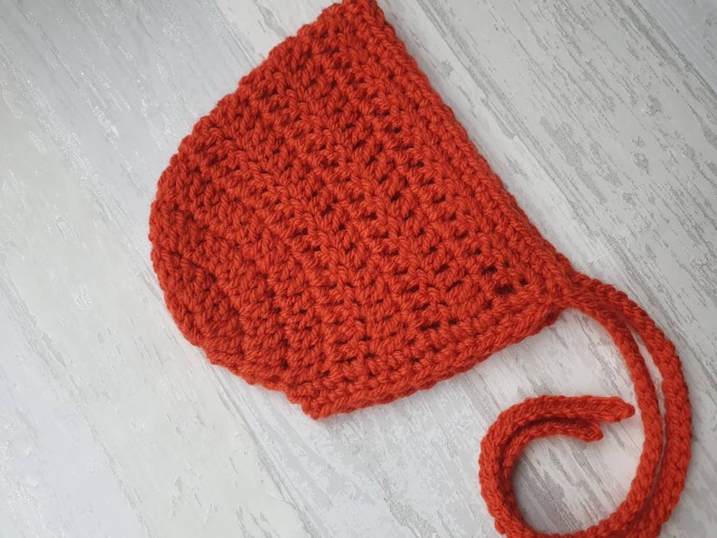 May include: A red crocheted baby bonnet with a long tie. The bonnet is made with a simple stitch pattern and has a soft, cozy feel.
