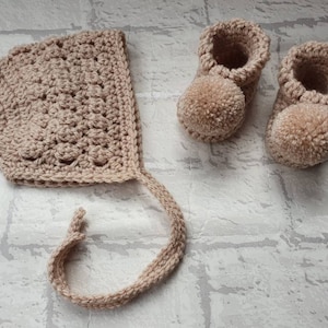 May include: A set of crocheted baby booties and a bonnet in a light beige color. The booties have pom-poms on the toes.