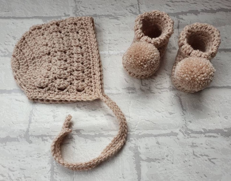 May include: A set of crocheted baby booties and a bonnet in a light beige color. The booties have pom-poms on the toes.