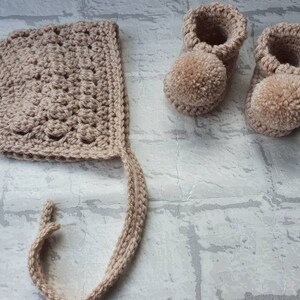 May include: A set of crocheted baby booties and a bonnet in a light beige color. The booties have pom-poms on the toes.