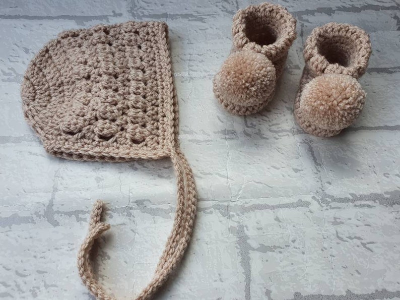 May include: A set of crocheted baby booties and a bonnet in a light beige color. The booties have pom-poms on the toes.
