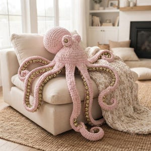 Könnte beinhalten: Ein großes, handgefertigtes Häkel-Oktopus-Stofftier aus weichem, pastellrosa Garn. Der Oktopus hat einen abgerundeten Kopf und acht lange, sich kräuselnde Tentakel. Die Unterseite jedes Tentakels ist mit einem schimmernden, goldfarbenen Paillettenstoff ausgekleidet.