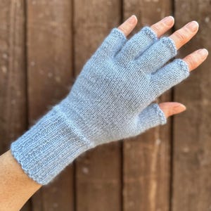 May include: Light blue fingerless gloves, knitted with a soft, textured yarn. The gloves have a ribbed cuff and are designed to leave the fingertips exposed. The background is a blurred wooden surface.