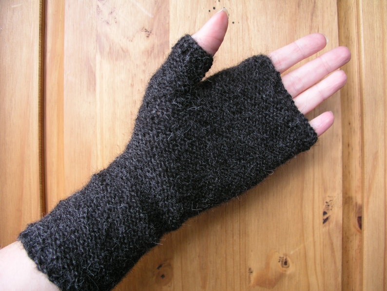 May include: A dark gray knitted fingerless glove, with a textured appearance. The glove covers the hand and wrist, leaving the fingers exposed. The knitted pattern is visible, and the material appears soft. The background is a light brown wooden surface.