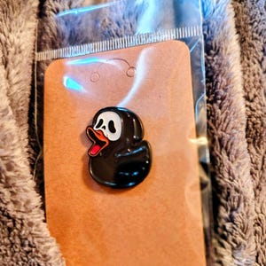 May include: A black enamel pin shaped like a rubber duck with a white ghost mask face, featuring a black hood, a bright orange beak, and a red interior mouth. The pin is attached to a rectangular brown cardboard backing inside a clear plastic sleeve.