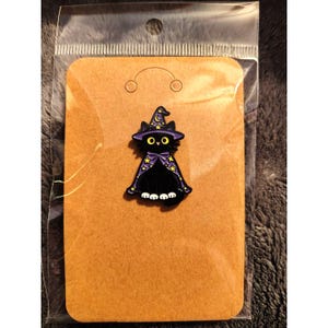 Puede incluir: Un pin de esmalte de gato negro con un sombrero de mago y una capa de color púrpura decorados con estrellas amarillas y una luna creciente. El gato tiene grandes ojos amarillos y patas blancas sobre una tarjeta de cartón marrón.