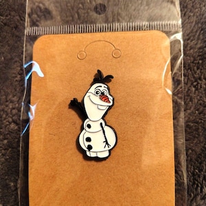 May include: A white snowman enamel pin with black outlines, a bright orange carrot nose, and three black buttons on the body. The snowman has a wide, friendly smile, stick-like arms, and small twigs on its head, presented on a brown card.