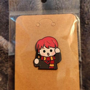 Puede incluir: Un pin de esmalte metálico que muestra un personaje de dibujos animados con cabello rojo brillante, vistiendo una túnica gris oscuro y una bufanda a rayas rojas y amarillas. El personaje sostiene una varita negra y tiene expresión de sorpresa.
