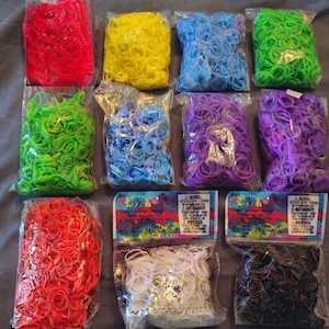 May include: A collection of eleven clear plastic bags filled with small, colorful rubber bands for crafting. The assortment includes red, yellow, light blue, green, purple, orange, white, and black bands, with two bags featuring Rainbow Loom labels.
